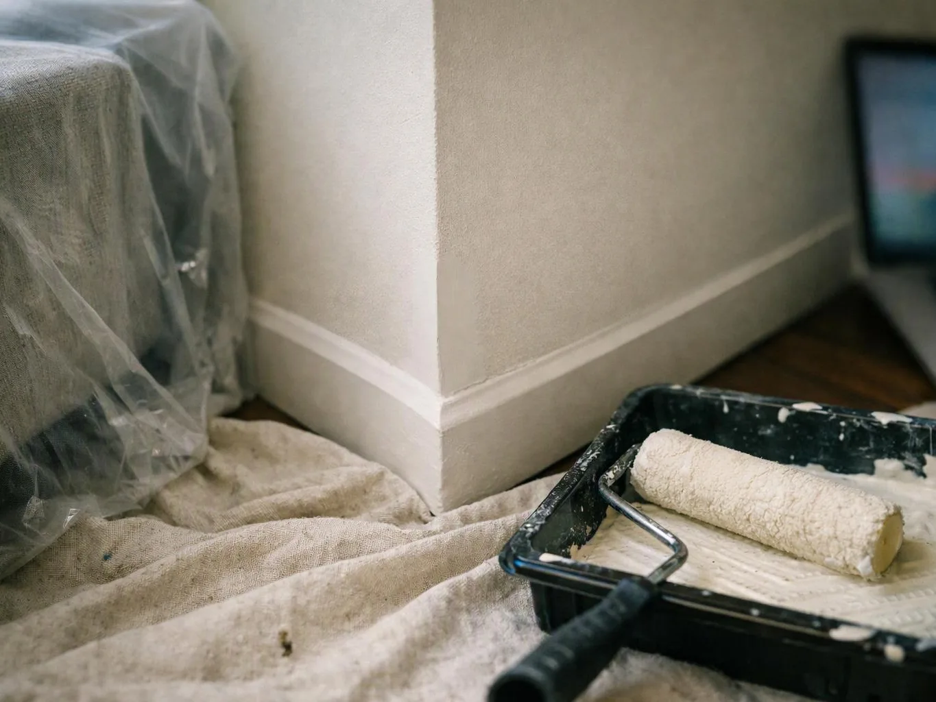 Paint tray with roller sits on a drop cloth beside a masked wall corner in a room under painting