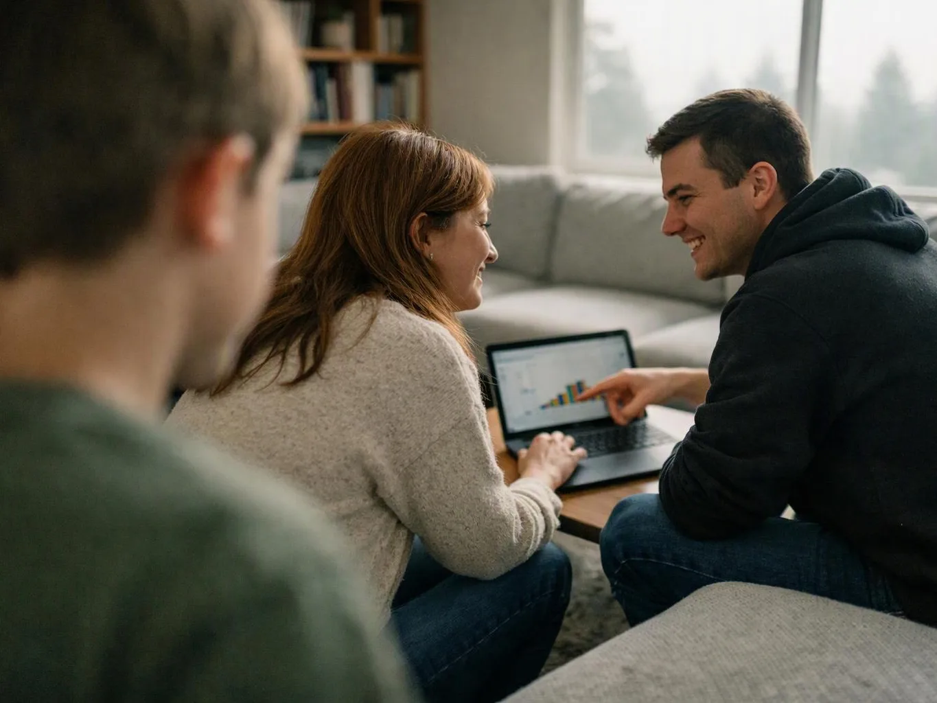 Two people smile at a laptop showing a colorful chart while a blurred person watches in a bright living room